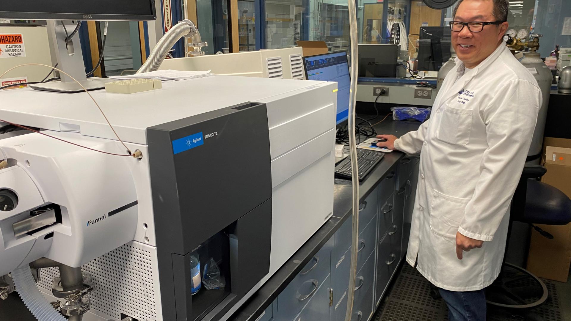 An analyst in a white lab coat stands to the right of an advanced laboratory instrument.