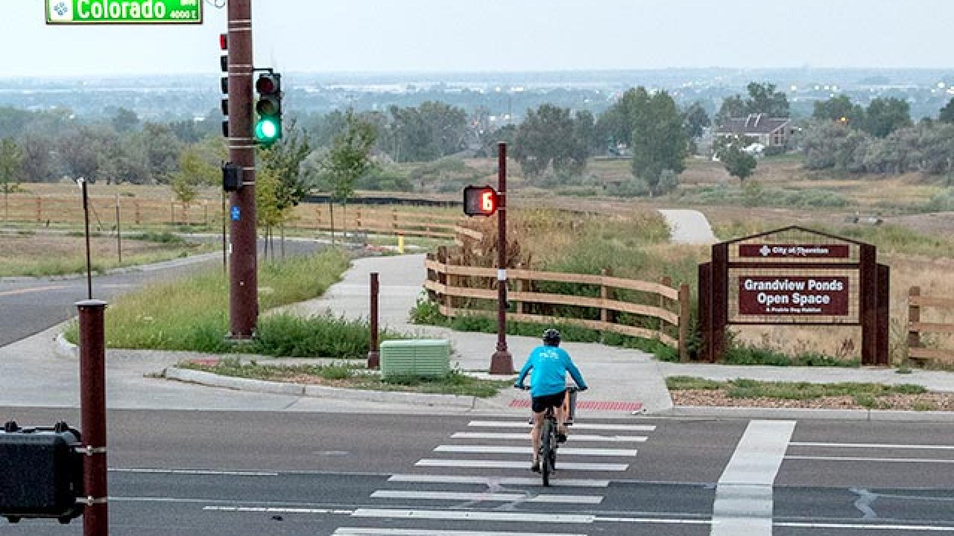 A cyclist wearing a blue jacket rides across a marked crosswalk at a signalized intersection towards the Grandview Ponds Open Space entrance on Colorado Boulevard south of 104th Avenue. A large city of Thornton sign to the right marks the entrance to “Grandview Ponds Open Space.” A paved path and wooden fence lead into a grassy, open landscape with scattered trees.