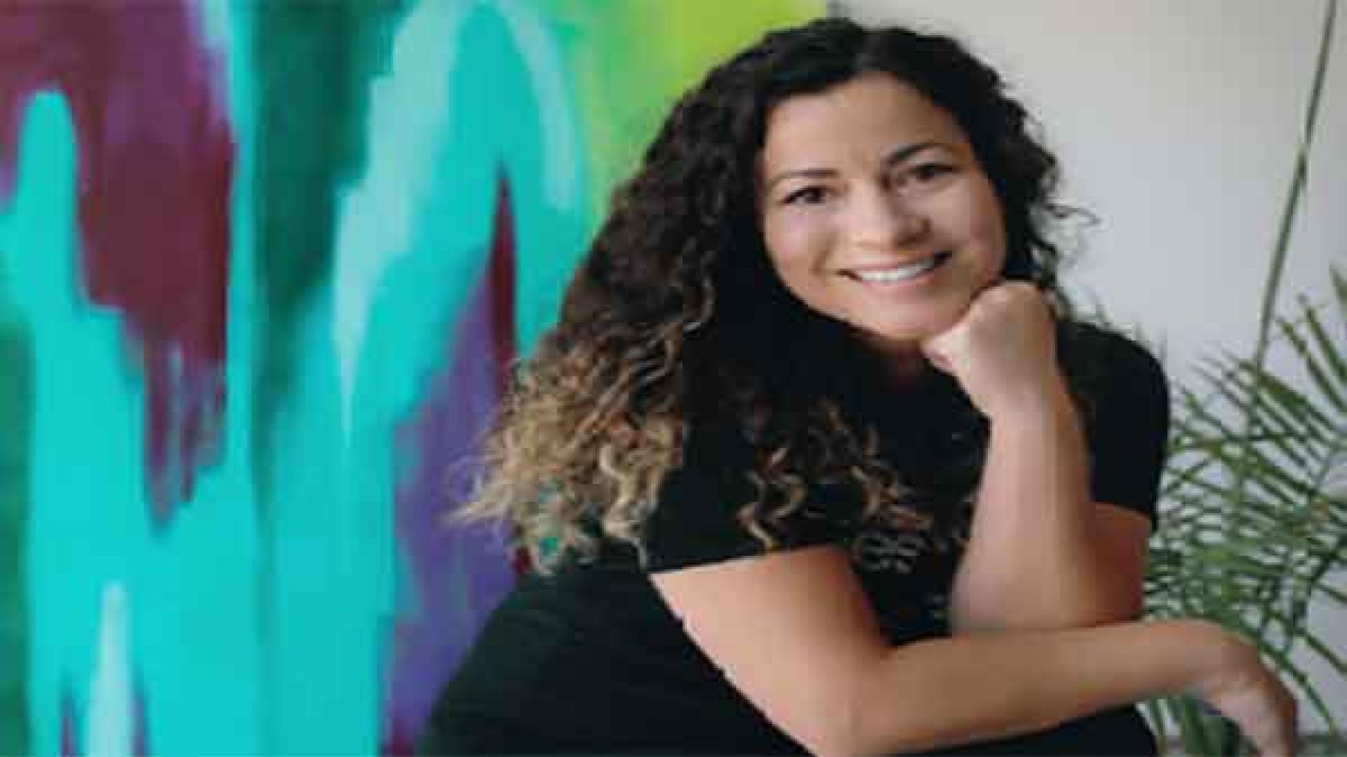 Latina artist seated in front of multicolor painting on wall.