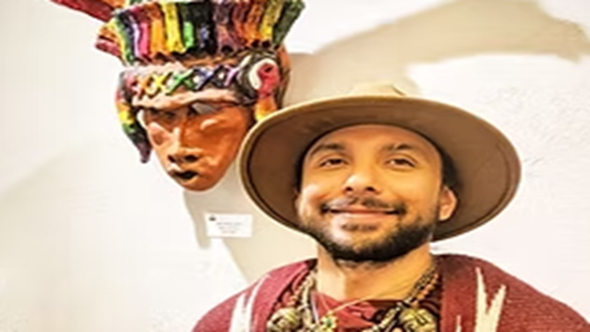 Hispanic artist wearing hat and standing in front of indigenous mask