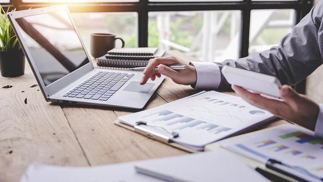 Laptop, tax forms, on a table and a man holding a calculator.