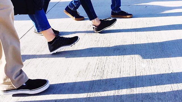 Close up of shoes and five people taking steps forward with their leg shadows cast on the concrete floor.