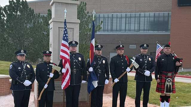 Seven-member honor guard in dark dress uniforms holding American and Colorado flags and ceremonial axes in front of an eagle-topped stone memorial at the Veterans Memorial Aquatic Center, with a bagpiper at the end of the formation.