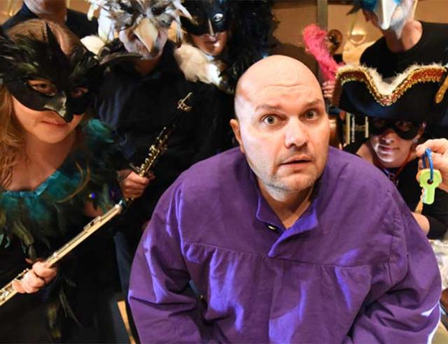 Actor in purple shirt surrounded by masked actors.