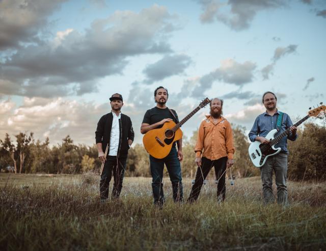 Four musicians stand in a grassy field at dusk, posing with their instruments including an acoustic guitar, bass guitar, and percussion. Trees and a cloudy sky fill the background, creating a relaxed outdoor band portrait.