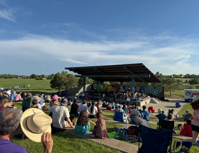 A lively outdoor concert in the park with a large crowd gathered on the grass, facing a stage set up at the front for live music. 