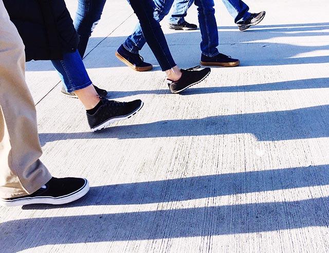 Close up of shoes and five people taking steps forward with their leg shadows cast on the concrete floor.