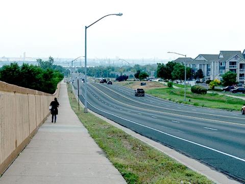 A person walks along a wide sidewalk beside a section of Colorado Boulevard with two lanes in each direction, a center turn lane, and bicycle lanes on both sides. A tall wooden fence lines the left side, while a grass buffer with streetlights separates the sidewalk from the road. The vehicle traffic is light with only a few cars traveling towards and away from the pedestrian in the distance.